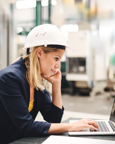 A side view of industrial woman engineer in a factory using laptop and smartphone.