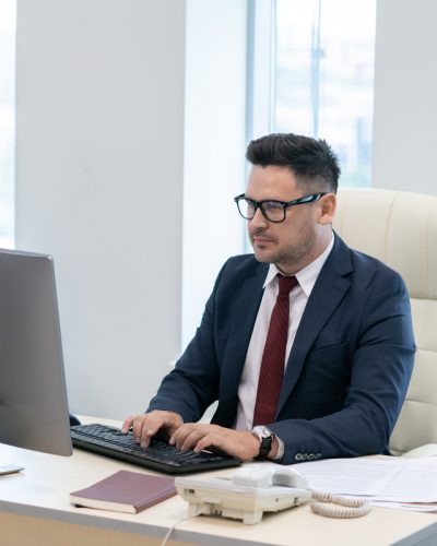 Busy director in elegant suit typing and looking at computer screen while sitting by desk in office